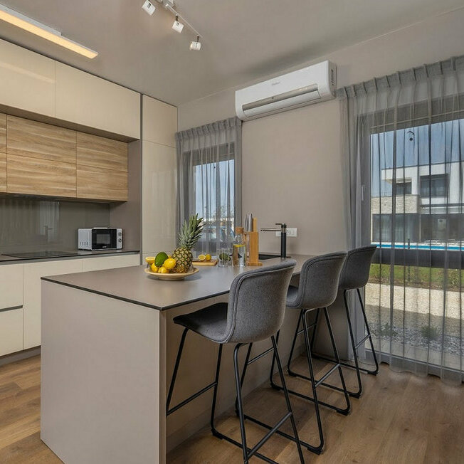 Moderna kuhinja s prostranim kuhinjskim otokom u središtu, savršenim za pripremu jela i druženje. Pregradne letvice diskretno odvajaju prostor, dodajući profinjen estetski dojam i toplinu interijeru.