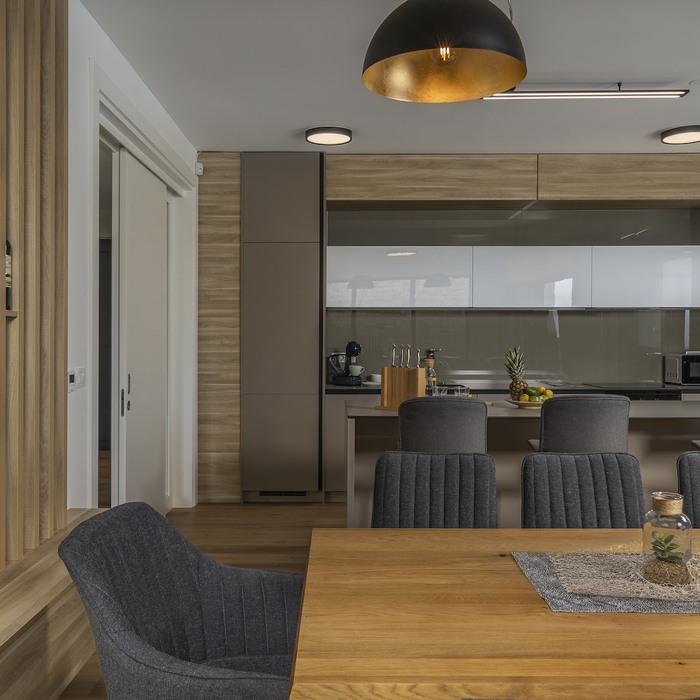 Ova moderna kuhinja ističe se elegantnim ugradbenim elementima u kombinaciji drvenih i neutralnih tonova, dok pregradbeni otok pruža funkcionalan i estetski dodatak prostoru. Pregradbene letvice diskretno razdvajaju blagovaonicu od dnevnog boravka, dodajući profinjenost i toplinu interijeru. Viseća komoda s bijelim frontama i zrcalnom površinom reflektira svjetlost, čineći prostor svjetlijim i vizualno prostranijim.